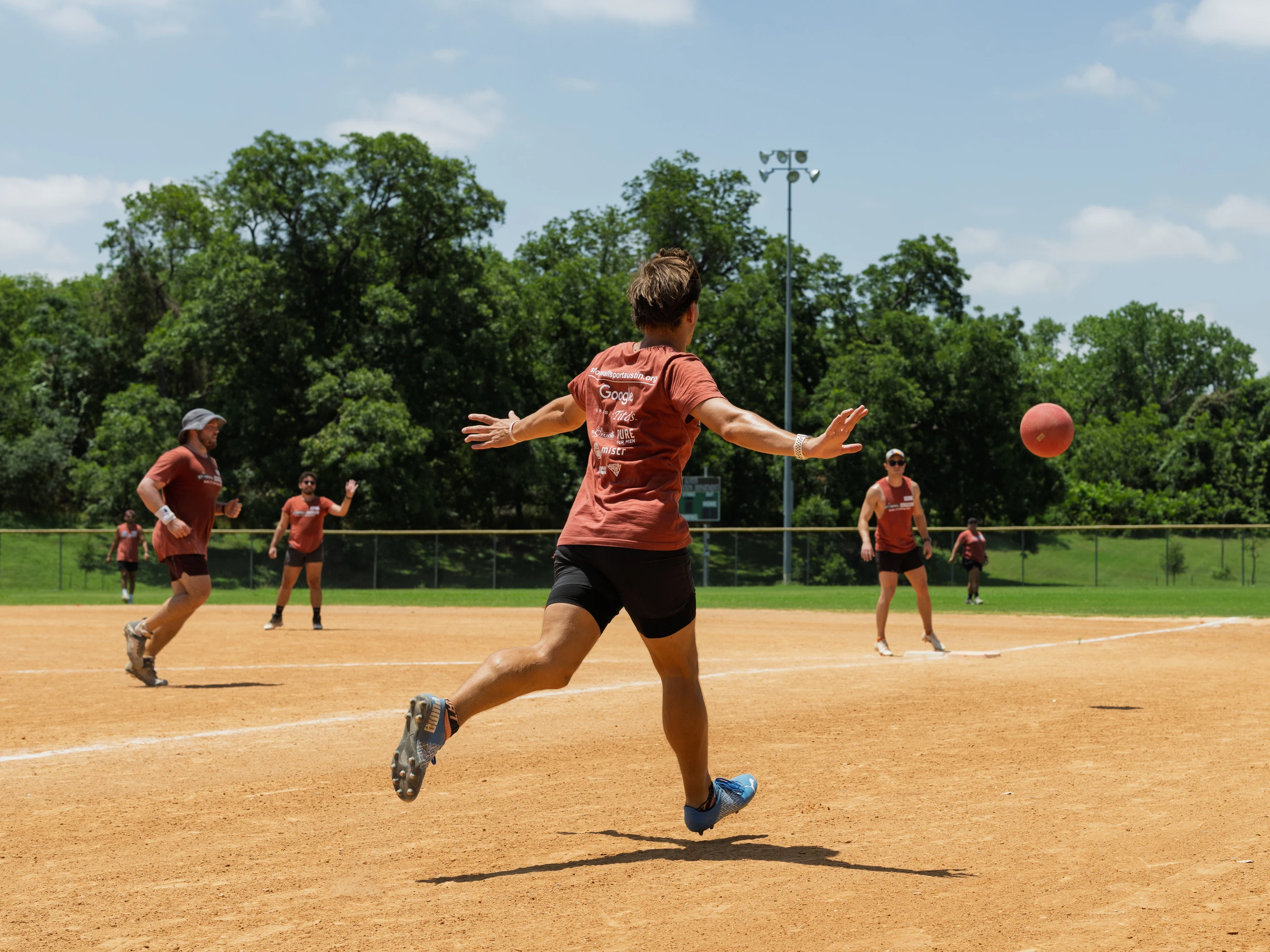 A player wearing a red team shirt and black shorts is in mid-stride after kicking a red kickball on a dirt kickball field, with several other players in similar uniforms positioned around the field under a sunny sky.