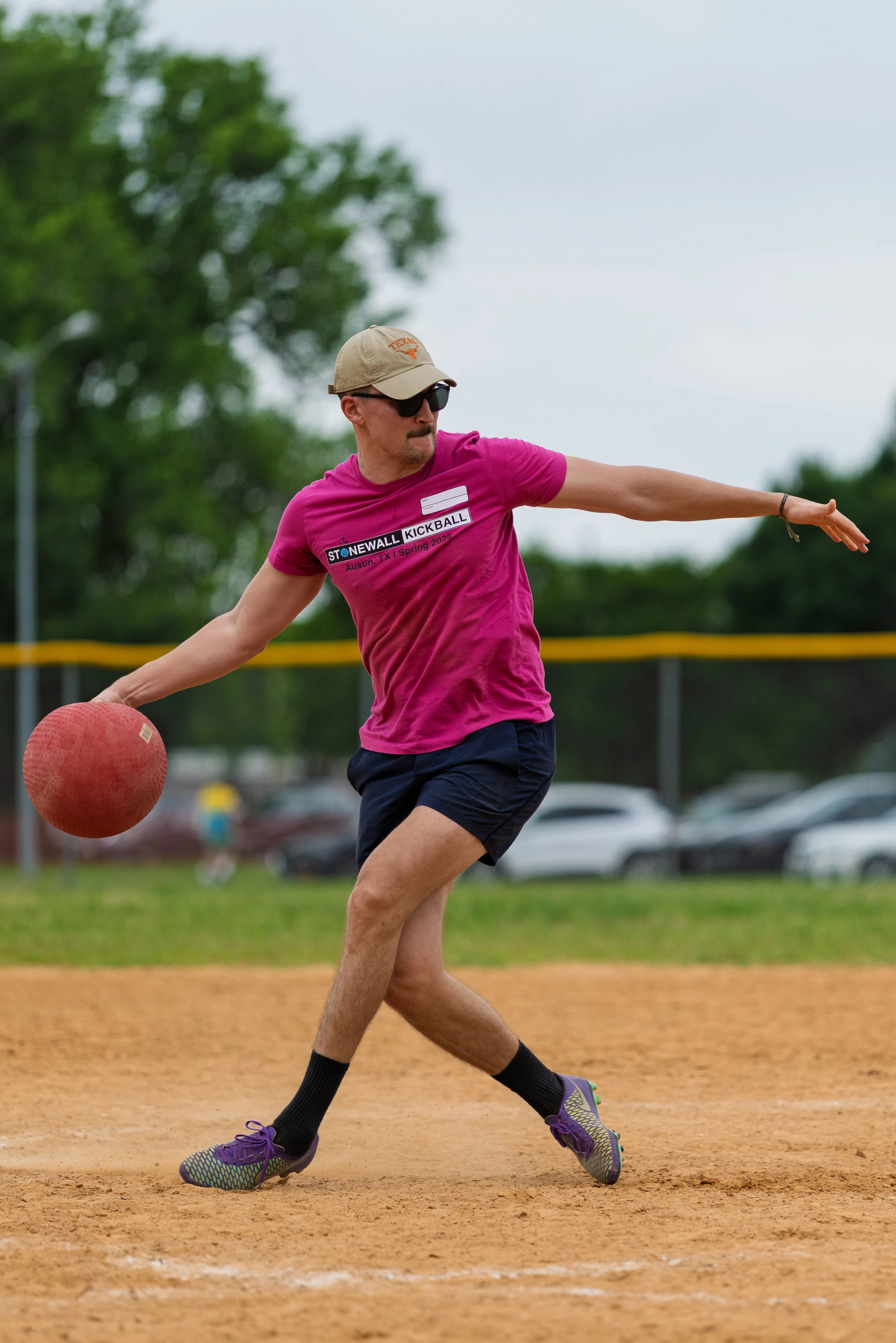 A player wearing a bright pink "Stonewall Kickball" shirt, shorts, a cap, and sunglasses is in the process of pitching a red kickball on a dirt field, with trees and a fence in the background.