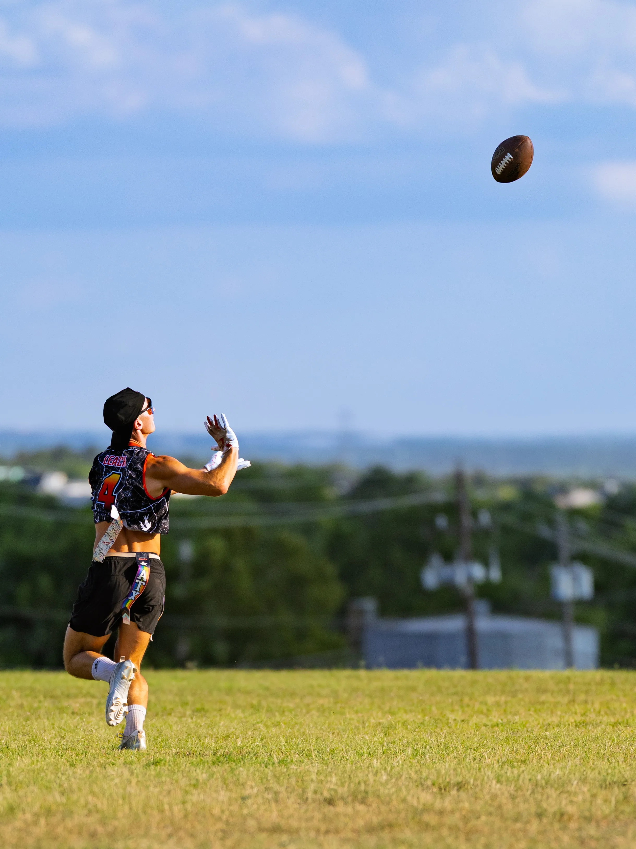 On a wide grassy field, an ATX Heat (Black) player in a black patterned crop jersey with the number 4, white gloves, cleats, a headwrap, and a flag belt sprints under a high pass, hands ready to catch the football; trees and utility lines line the distant horizon under a blue sky.