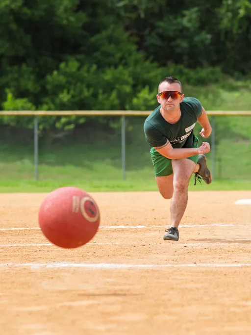 A player wearing a green shirt, green shorts, and red sunglasses is in motion, having just rolled a red kickball across a dirt field, with green grass and trees visible in the background.
