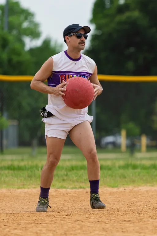 A kickball player wearing a white and purple uniform, purple socks, athletic shoes, a black cap, and sunglasses is standing on a dirt field while holding a red kickball, appearing focused and ready to make a play. Trees and a fence are visible in the background.