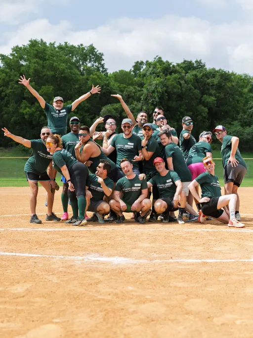 A group of kickball players pose energetically on a sandy kickball field, wearing matching green team shirts and a variety of athletic shorts, hats, and sunglasses, with trees visible in the background.