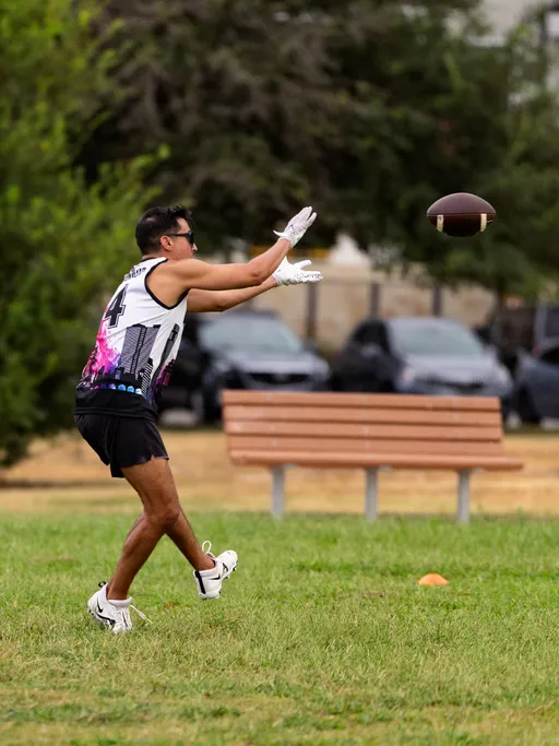 An ATX Heat (Orange) player in a sleeveless white-and-black jersey with the number 4, black shorts, white cleats, gloves, and sunglasses reaches forward to catch a football during practice drills on a grassy park field. An orange cone marks the area, with a bench and parked cars in the background.