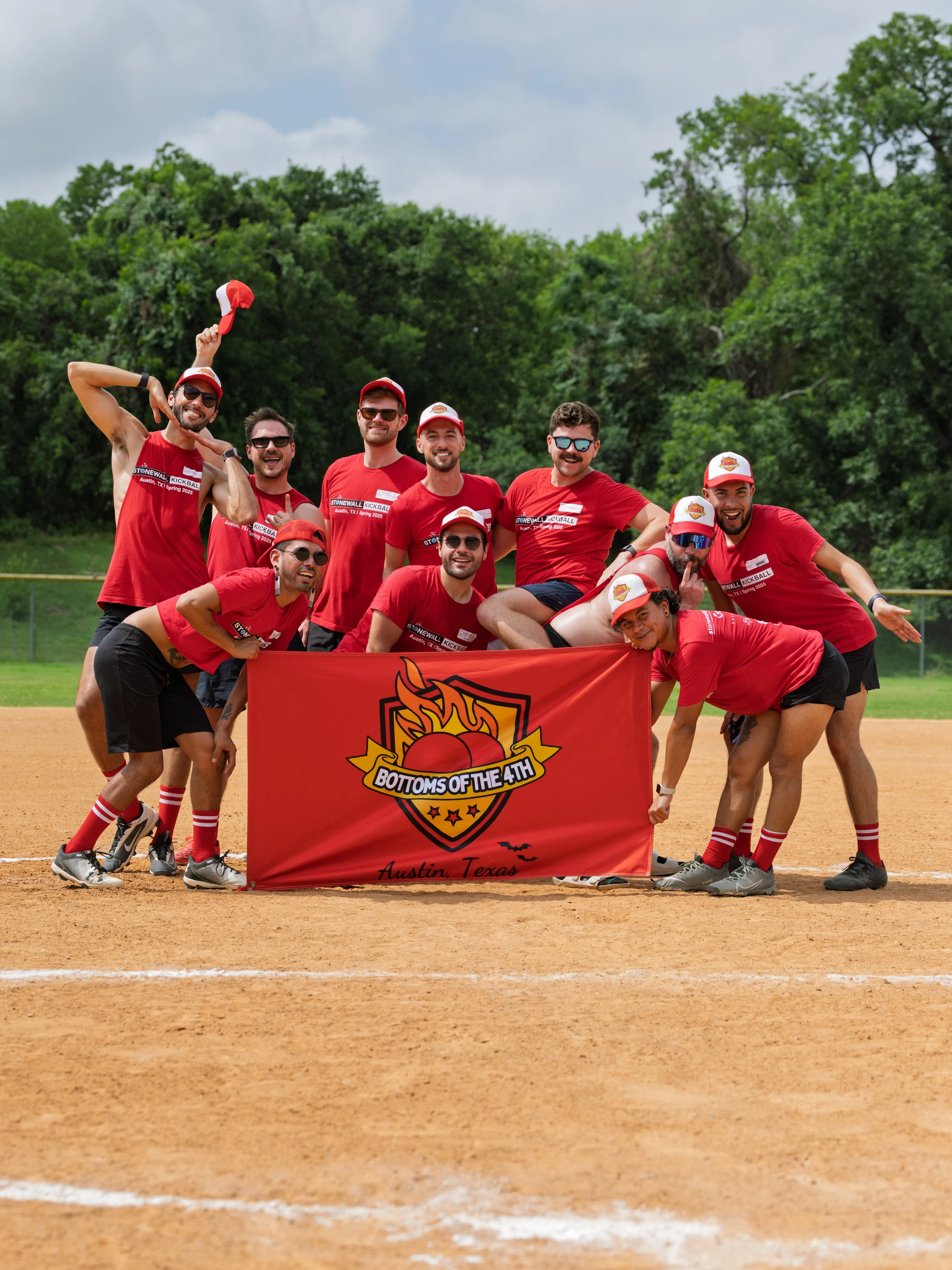 A group of kickball players poses together on a kickball field, all wearing matching red shirts, hats, and socks, smiling and gathering around a team banner that reads "Bottoms of the 4th, Austin, Texas."