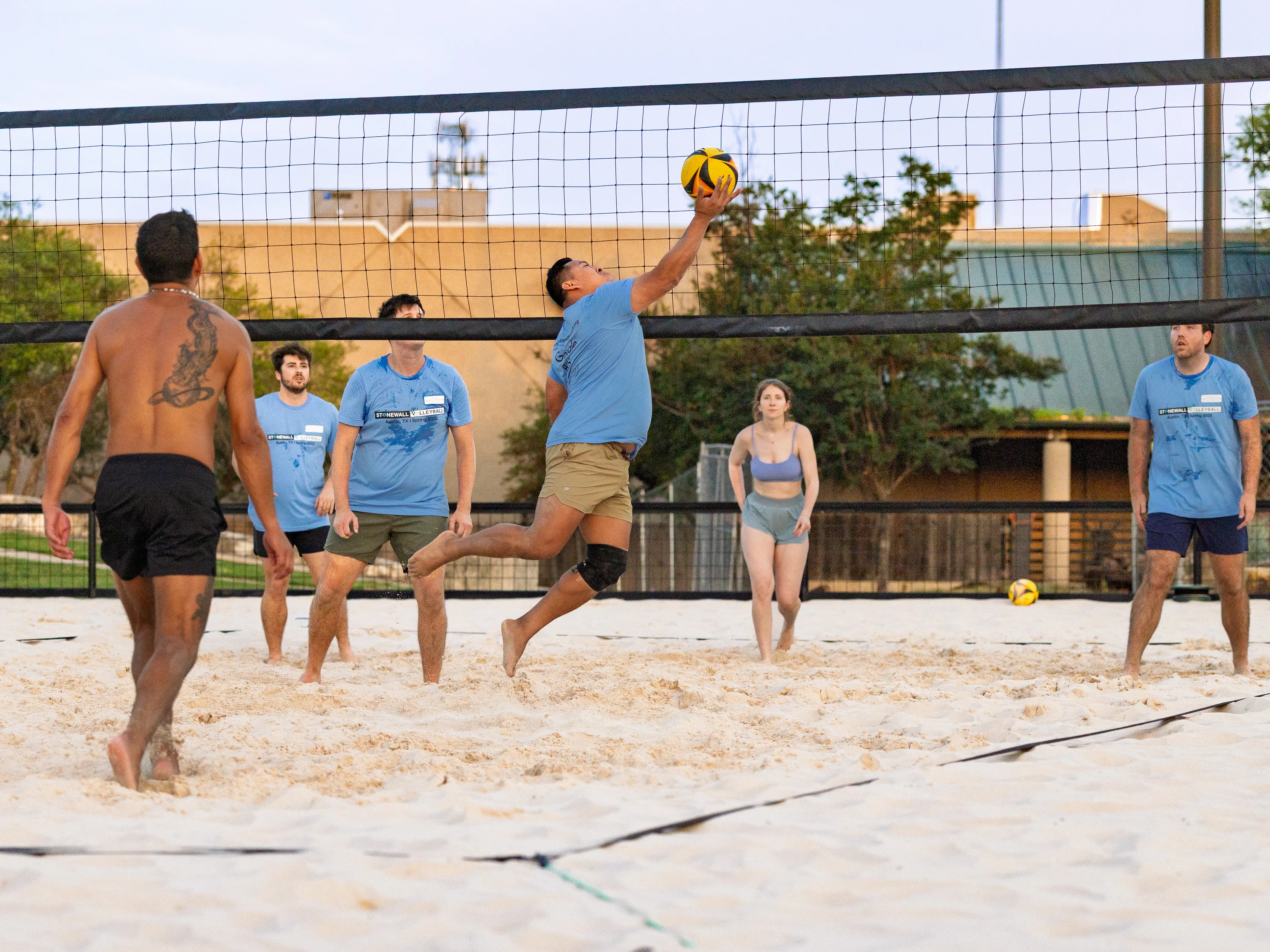 On a sand volleyball court, a player in a blue team shirt and knee brace leaps at the net to tap a yellow-and-black volleyball, while teammates in matching shirts ready behind. Opponents on the near side watch the play amid park trees and buildings.
