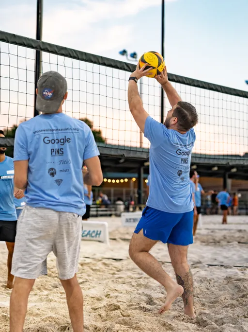 Two volleyball players wearing matching light blue shirts are on a sand court, with one player jumping and raising their arms to set a volleyball near the net while the other stands nearby watching. The game takes place outdoors as evening approaches, with several people and equipment visible in the background.