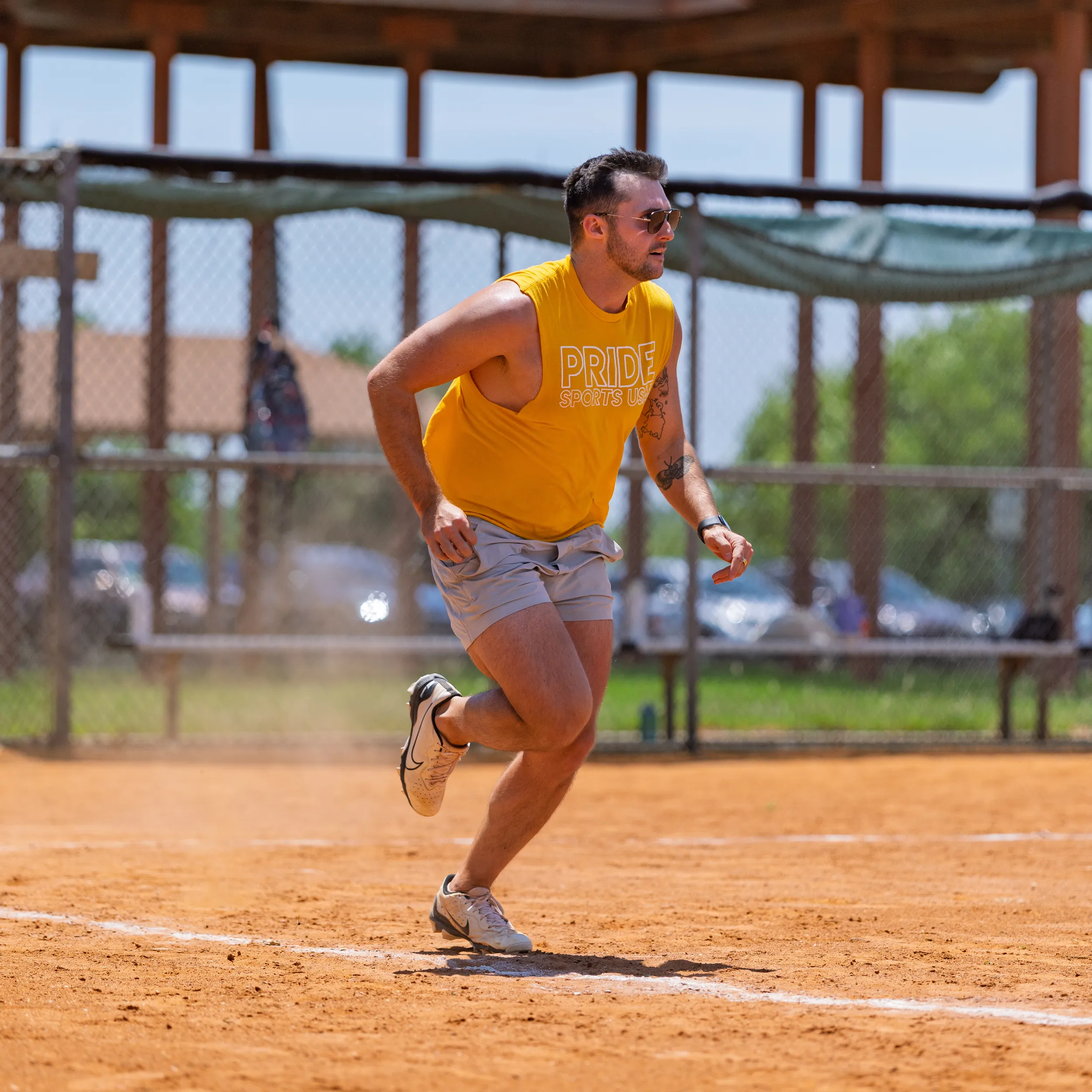 A player in a yellow PRIDE SPORTS tank, gray shorts, sunglasses, and running shoes sprints up the baseline on a dusty kickball field, dirt kicking up behind. Chain-link fencing and shaded benches line the background.