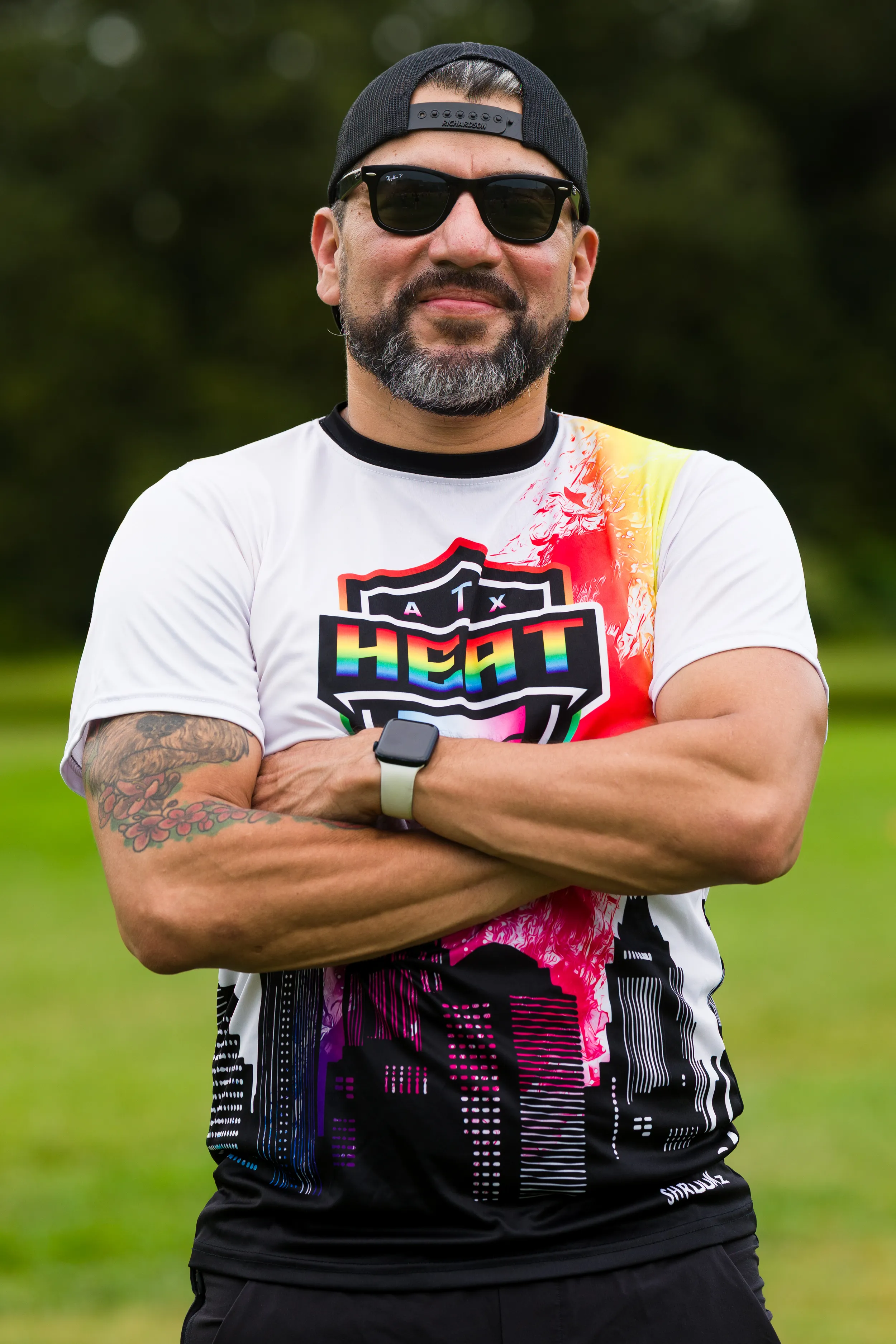 On a grassy practice field, an ATX Heat (Orange) player stands with arms crossed during Gay Bowl 2025 prep, wearing a white-and-orange ATX Heat jersey with a rainbow-shield logo and city-skyline graphic, black shorts, sunglasses, a backward cap, a smartwatch, and a visible forearm tattoo.