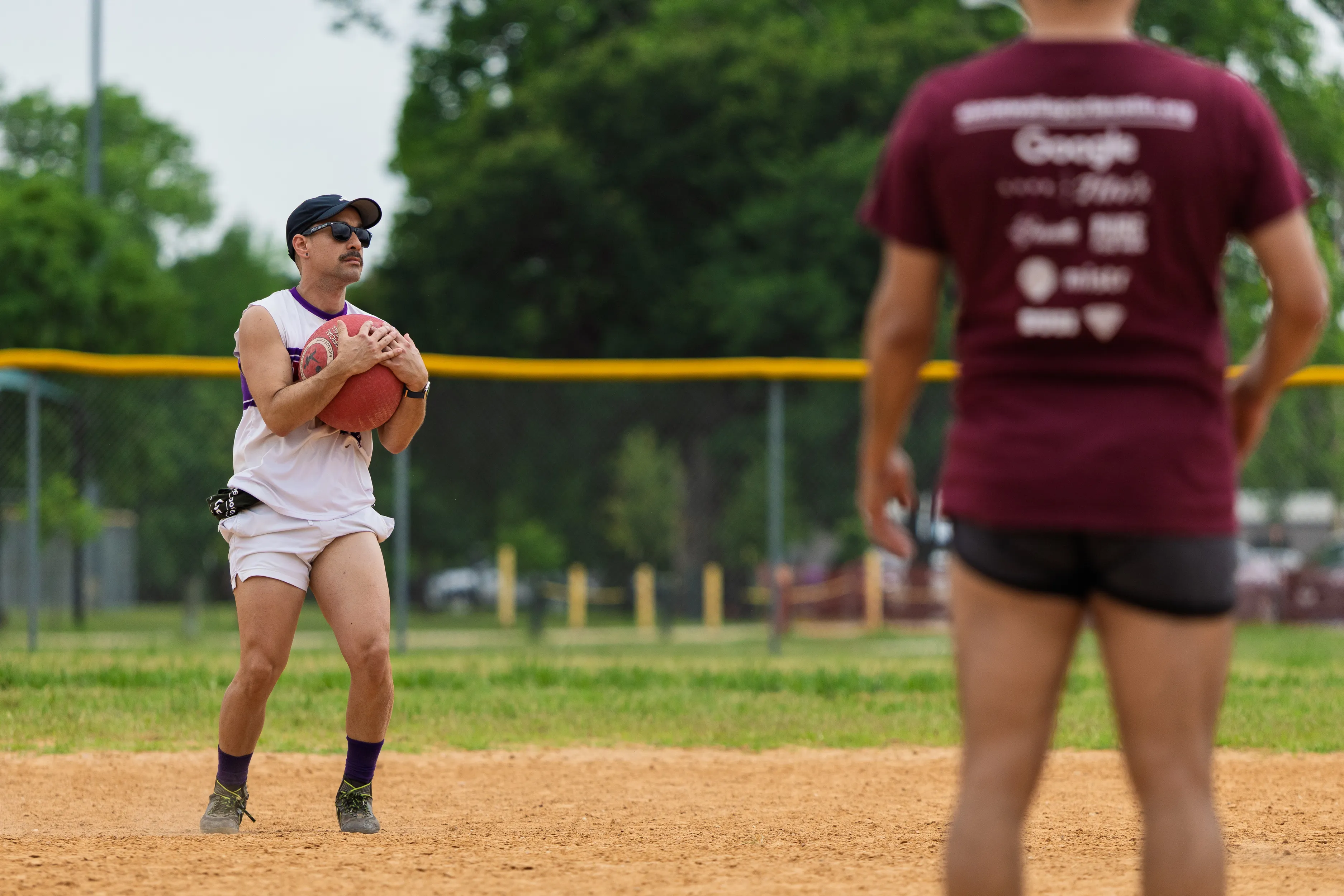 On a dirt kickball field, a player in a white-and-purple uniform with knee-high purple socks, sunglasses, and a black cap cradles a red kickball, looking ready to throw. Another player in a maroon shirt and black shorts stands in the foreground, with a yellow outfield fence and trees in the background.
