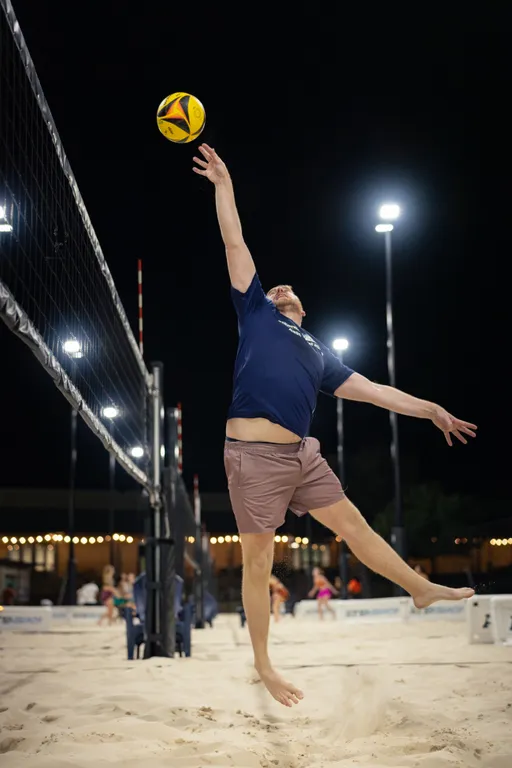 On a lit sand volleyball court at night, a player in a navy shirt and mauve shorts jumps barefoot beside the net, reaching one-handed toward a yellow-and-blue ball for a tip or spike. String lights, tall poles, and other courts with players appear in the background.