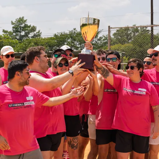 A group of kickball players wearing matching bright pink "Stonewall Kickball" shirts is gathered on a kickball field, cheering and raising a large trophy together in celebration.