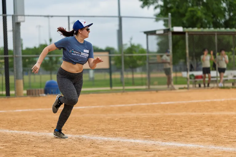 A player wearing a Stonewall Kickball shirt, leggings, and a baseball cap is running on a kickball field, with teammates and a dugout visible in the background.