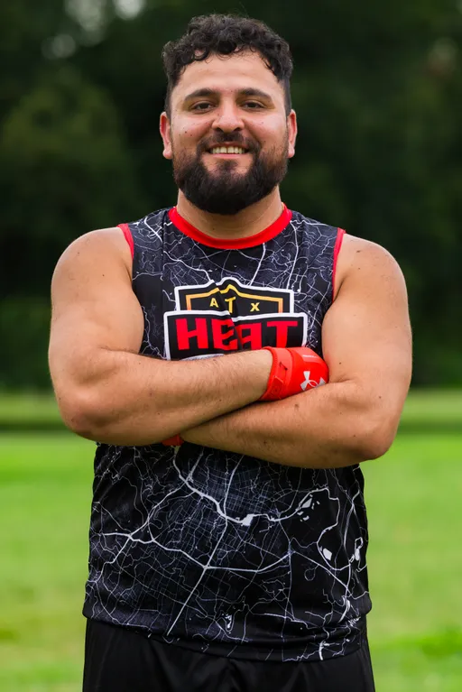 An ATX Heat (Orange) player stands on a grassy field during practice for Gay Bowl 2025, arms crossed, wearing a black map-pattern sleeveless jersey with red trim and an “ATX HEAT” logo, plus bright red receiver gloves, with trees blurred behind.