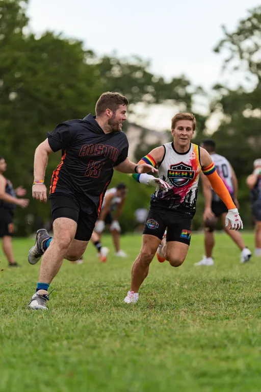 Two ATX Heat (Orange) players sprint during flag football practice for Gay Bowl 2025, with one in a black “AUSTIN 13” kit running upfield while a defender in a white-and-orange sleeveless jersey, gloves, and an arm sleeve reaches to make a tag. Teammates move in the background on a grassy field bordered by trees.