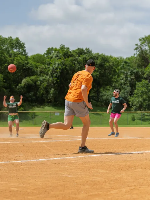 A player in an orange shirt and gray shorts is running across a kickball field while two other players in green shirts prepare to catch an airborne kickball, with trees and a fence in the background.