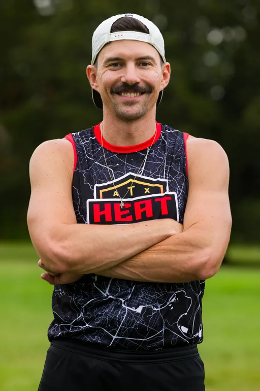 An ATX Heat player stands on a grassy field during team practice for Gay Bowl 2025, arms crossed and smiling, wearing a black sleeveless ATX Heat jersey with red trim and a map-like pattern, a backward white cap, and a small pendant necklace; blurred trees fill the background.
