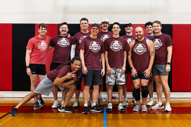 A dodgeball team poses on an indoor gym court in front of a red-and-black padded wall, with players standing and crouching in a relaxed group. Most wear matching maroon “Texas Premier” shirts with athletic shorts and sneakers; a few have a headband, baseball cap, or knee brace, and one player leans forward playfully at the front.