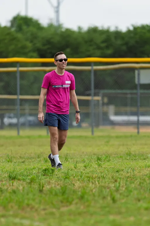 A player wearing a bright pink "Stonewall Kickball" shirt, dark shorts, athletic shoes, white socks, and sunglasses is walking on a grassy kickball field, with a chain-link fence and trees in the background.
