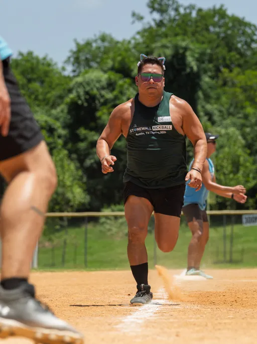 A player wearing a dark tank top with "Stonewall Kickball" text, shorts, black sneakers, sunglasses, and cat ear headbands is running on a kickball field with other players visible in the background. The scene is outdoors on a sunny day with green trees behind the field.