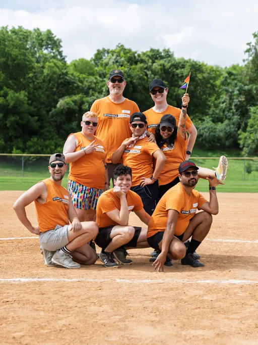 A group of eight kickball players pose together on a dirt field, all wearing matching orange "Stonewall Kickball" shirts; one person holds a small rainbow flag, and some players are kneeling or crouching in front while others stand behind with trees and greenery in the background.