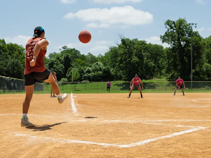 A player in athletic wear is kicking a red kickball on a sandy outdoor field, while several other players in matching shirts prepare defensively in the background under a bright sky.