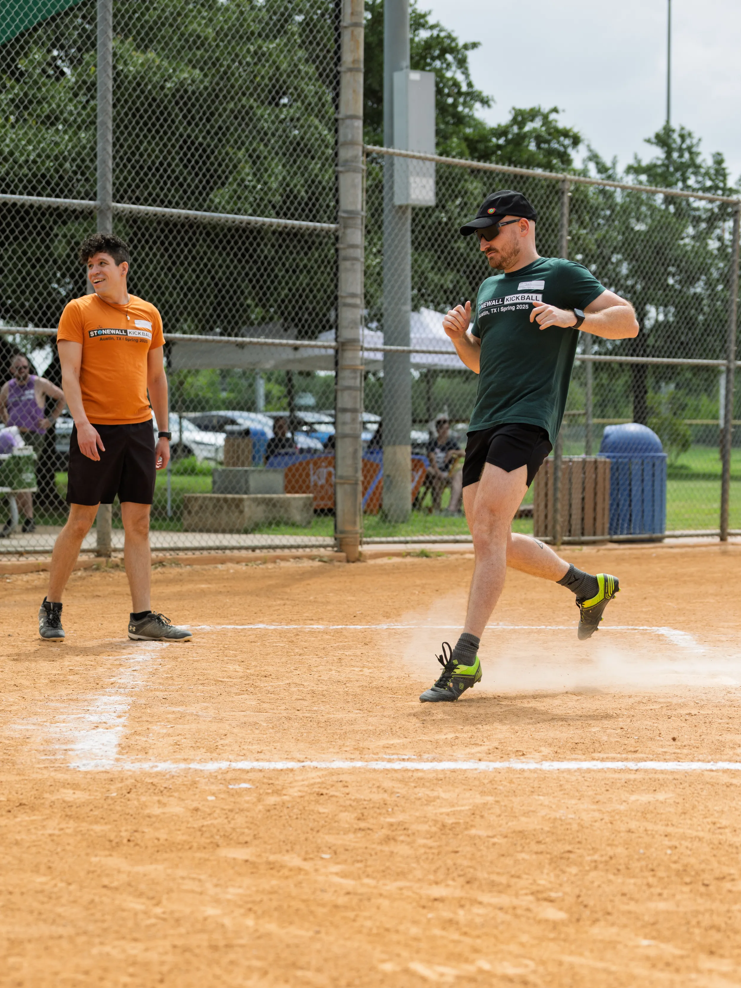 A player in a green Stonewall Kickball shirt, black shorts, cap, and sunglasses crosses home plate, kicking up dust on a kickball field, while a player in an orange team shirt watches nearby; chain-link fencing and park greenery frame the scene.