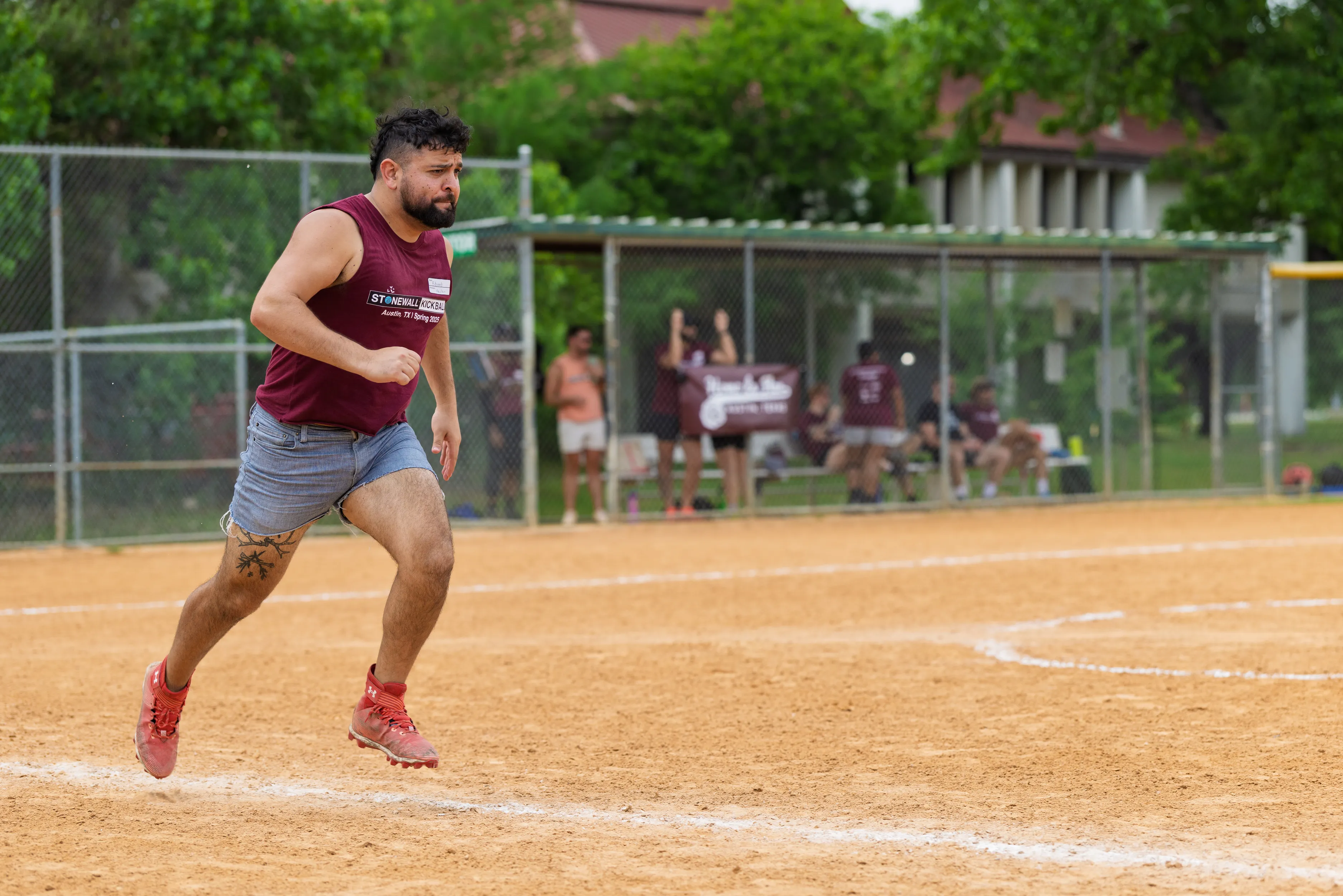 A player in a maroon Stonewall Kickball sleeveless jersey, gray shorts, and red cleats sprints across a dirt kickball field, captured mid-stride along the chalked baseline. Behind a chain-link fence, teammates in matching maroon shirts watch and cheer from the dugout area.