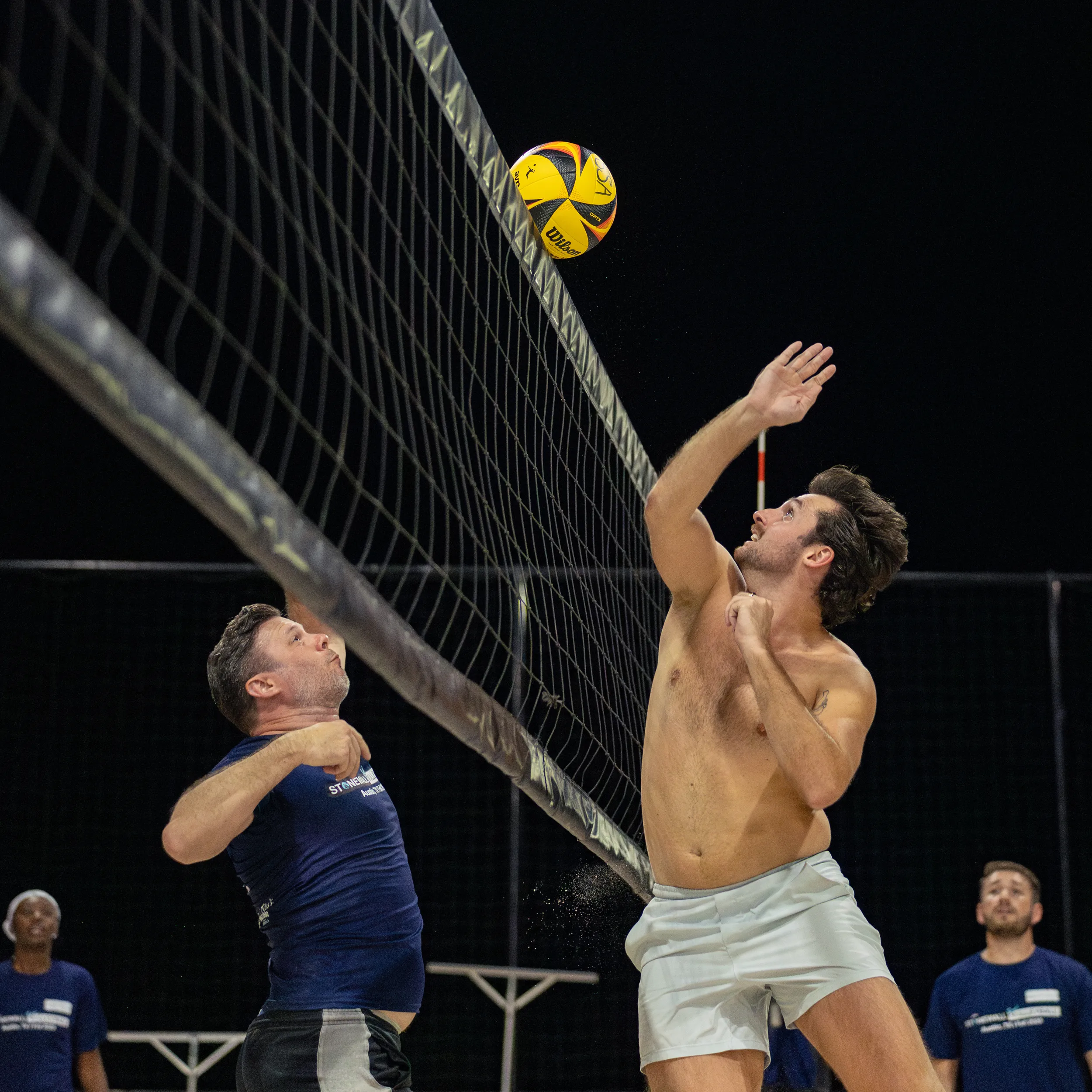 On a lit sand volleyball court at night, a player in white shorts jumps at the net to tip or block a yellow-and-black volleyball near the antenna while an opposing player in a navy shirt reaches up from the other side. Teammates in matching shirts watch from the background as the rally unfolds.