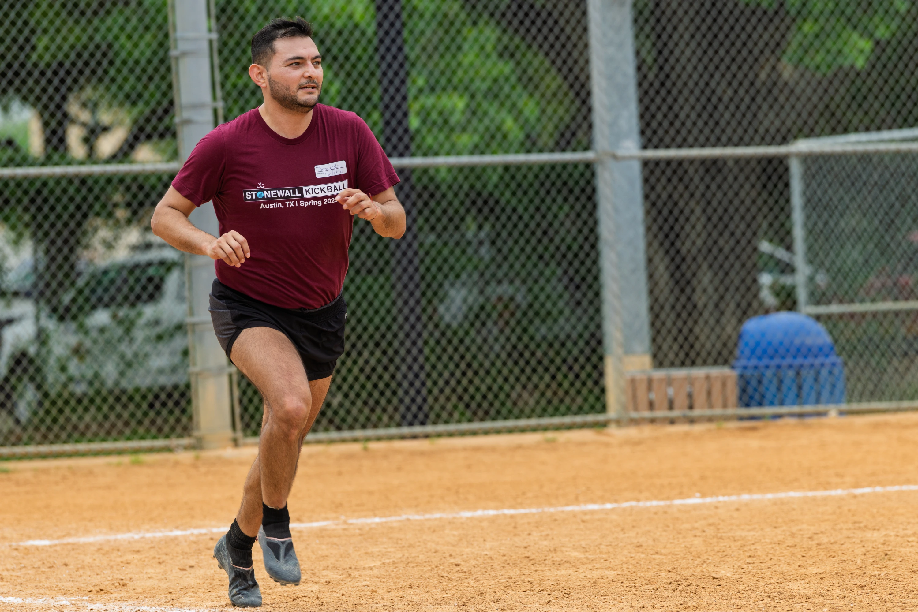 A player wearing a maroon "Stonewall Kickball" t-shirt and black shorts is running on a kickball field, with a chain-link fence and trees in the background.
