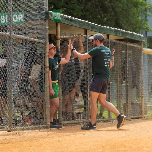 A player wearing a green shirt and shorts is giving a high five to another player standing in the dugout area next to the kickball field, with both individuals appearing cheerful and engaged in the game. The environment includes fencing, signage that reads "VISITOR", and other players and trees in the background.