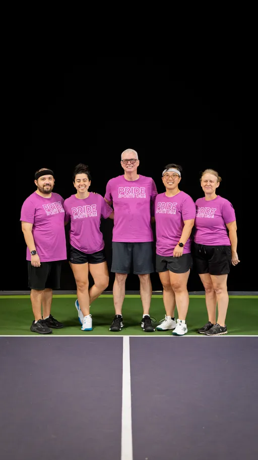 Five pickleball players stand together on an indoor court, all wearing matching pink "Pride Pickleball Summer 2025" shirts, shorts, and athletic footwear, some with headbands or glasses, posing for a group photo.