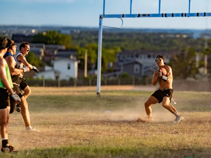 On a dusty grass field at sunset, an ATX Heat (Black) receiver secures a football near a goal frame while wearing black shorts, a headband, sunglasses, cleats, and a flag belt; two defenders in black practice gear and flag belts chase from the left, with suburban houses and hills in the background.