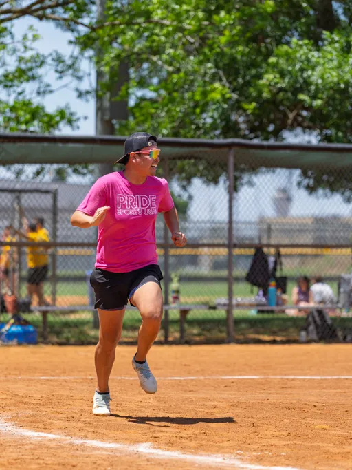 A player wearing a bright pink "Pride Sports USA" shirt, black shorts, a backwards cap, and sunglasses is running on a dusty kickball field, with a chain-link fence and trees in the background.