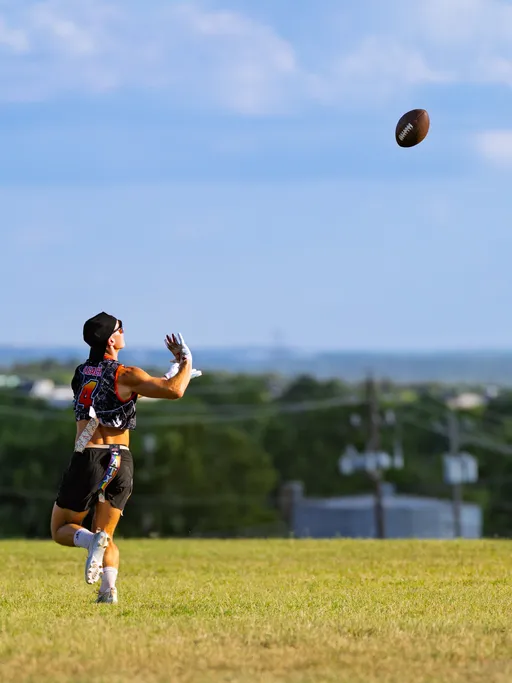 On a wide grassy field, an ATX Heat (Black) player in a black patterned crop jersey with the number 4, white gloves, cleats, a headwrap, and a flag belt sprints under a high pass, hands ready to catch the football; trees and utility lines line the distant horizon under a blue sky.