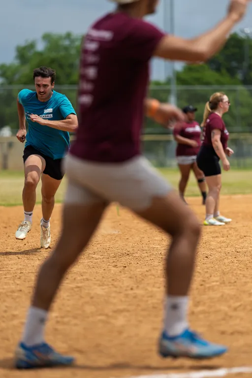 A player in a blue shirt sprints toward a base on a kickball field while another player in a maroon shirt and white shorts stands in the foreground; additional players are visible in the background, all appearing focused on the ongoing game.