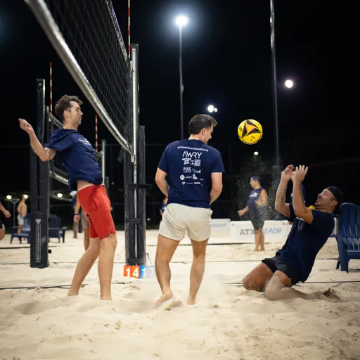 Night sand volleyball action: three players in navy shirts react as a yellow-and-black ball drops near the net—one jumping by the pole, one bracing in tan shorts, and one falling backward into the sand with hands up to make a play. The scene is lit for night play with multiple courts and blue chairs visible in the background.