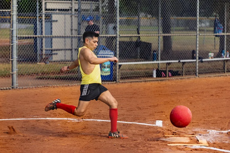 On a red-dirt kickball field, a player in a yellow tank top, black shorts, knee-high red socks, and cleats drives a red kickball off the home-plate line in mid-kick. A chain-link fence, dugout bench with gear, and chalked baselines frame the in-game moment.