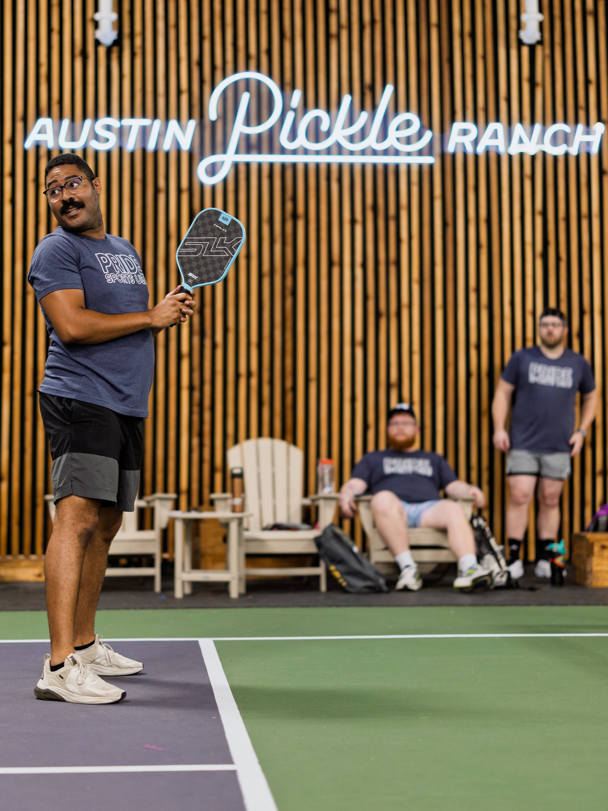 A player on a pickleball court prepares to swing a paddle, wearing athletic shorts, a "Pride Pickleball" t-shirt, and sneakers; in the background, two other participants in matching t-shirts observe from near wooden lounge chairs at the Austin Pickle Ranch indoor facility.