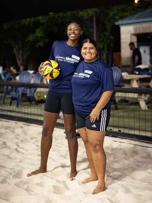 Two players stand barefoot on a lit sand volleyball court, smiling side by side. They wear navy Stonewall Volleyball shirts and black shorts; one holds a yellow-and-black volleyball, with fencing, benches, and lounge chairs in the background.