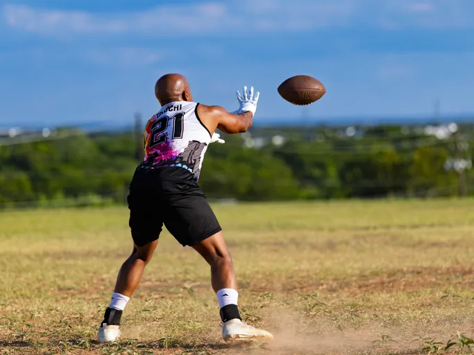 During ATX Heat (Black) practice for Gay Bowl 2025, a player wearing a sleeveless team jersey (number 21), black shorts, white gloves, and cleats reaches out to catch a football on an open grassy field with distant trees under a bright sky.