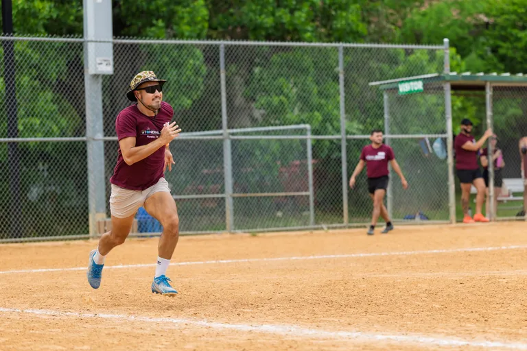 A kickball player wearing a maroon Stonewall team shirt, light shorts, blue cleats, sunglasses, and a bucket hat is running on the dirt field, with other team members and a chain-link fence visible in the background.