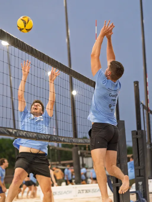 Two volleyball players in light blue shirts and black shorts are jumping at the net, one attempting to block while the other prepares to spike the ball on a sand volleyball court during a match, with other competitors visible in the background.