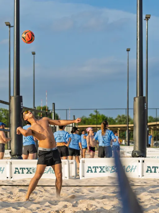 A volleyball player is preparing to serve the ball on a sand court, wearing black shorts, sunglasses, and a visor. Other athletes in blue shirts are gathered in the background, with sand, volleyball nets, and outdoor lighting visible around the court.