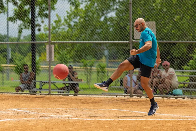 A kickball player wearing a teal shirt, black shorts, and athletic shoes is kicking a red kickball on a dirt field, with a chain-link fence and spectators seated on benches in the background.