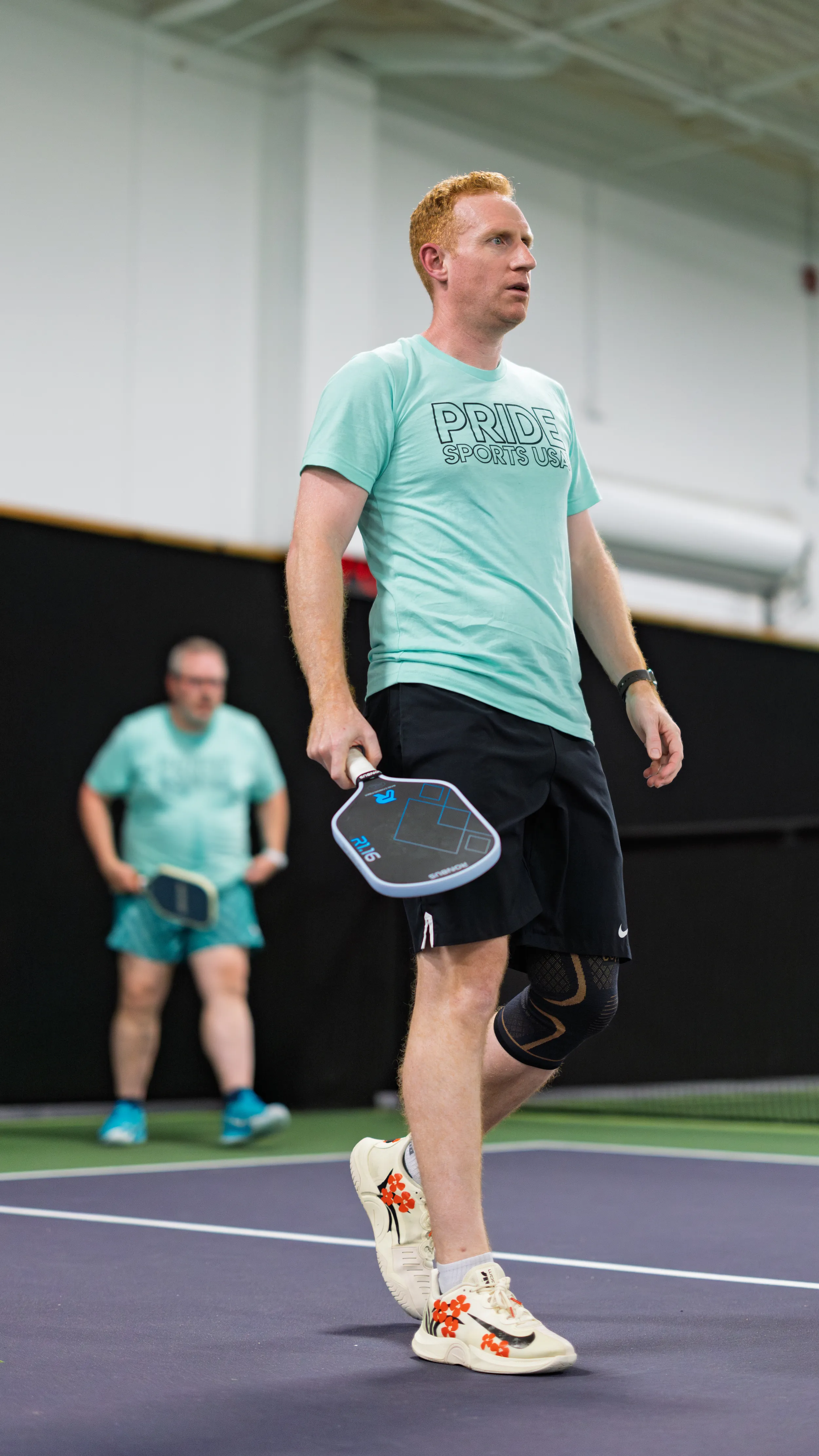 On an indoor pickleball court, a player in a mint “PRIDE SPORTS USA” shirt, black shorts, and floral sneakers steps forward holding a paddle, a knee brace visible on one leg. Another player in similar mint gear stands ready in the background on the purple-and-green court, awaiting the next rally.