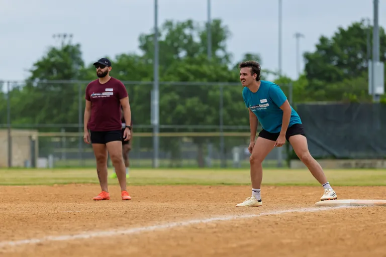 Two kickball players are on a dirt field; one athlete in a teal shirt and athletic shorts is crouched and ready near a base, while another player in a maroon shirt and shorts stands watching from further back. The setting is an outdoor kickball field with visible fences and green trees in the background.