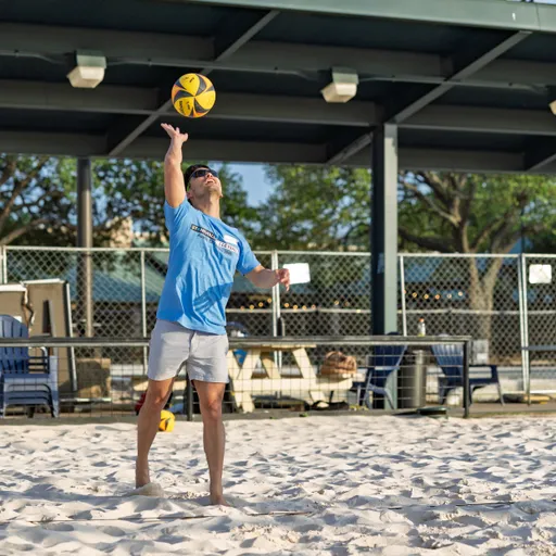A volleyball player wearing a light blue t-shirt and shorts is poised to serve, tossing a yellow and blue volleyball into the air on a sand court with outdoor seating and fencing visible in the background.