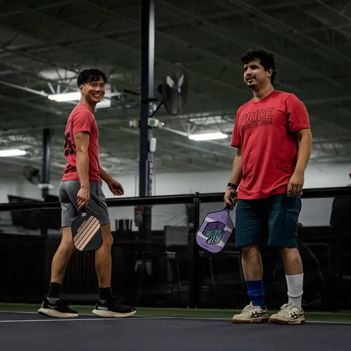 Two pickleball players stand on an indoor court, each holding a paddle and wearing athletic clothing, including red "PRIDE" shirts. They appear to be between points, with one player smiling toward the camera and a net visible behind them.