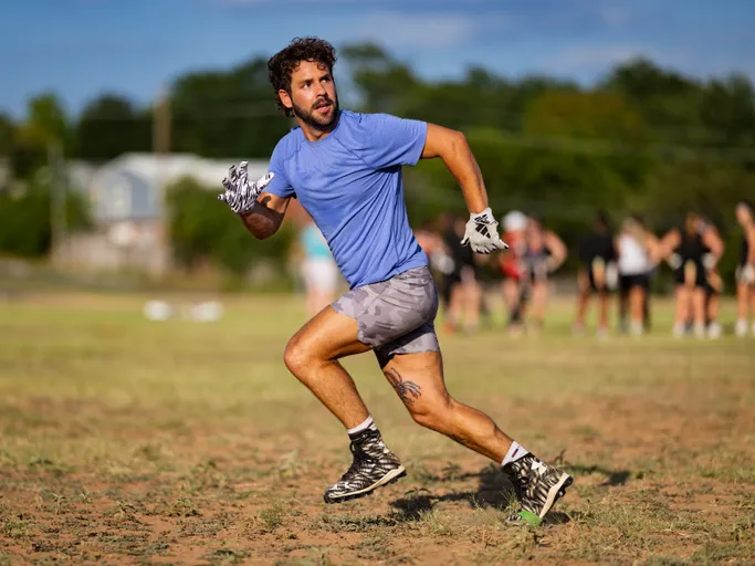 ATX Heat (Black) player sprints and makes a sharp cut during flag football practice on a dry grass field, eyes up and gloved hands ready. The athlete wears a blue shirt, gray shorts, and black cleats, with teammates blurred in the background near the sideline under a sunny sky.
