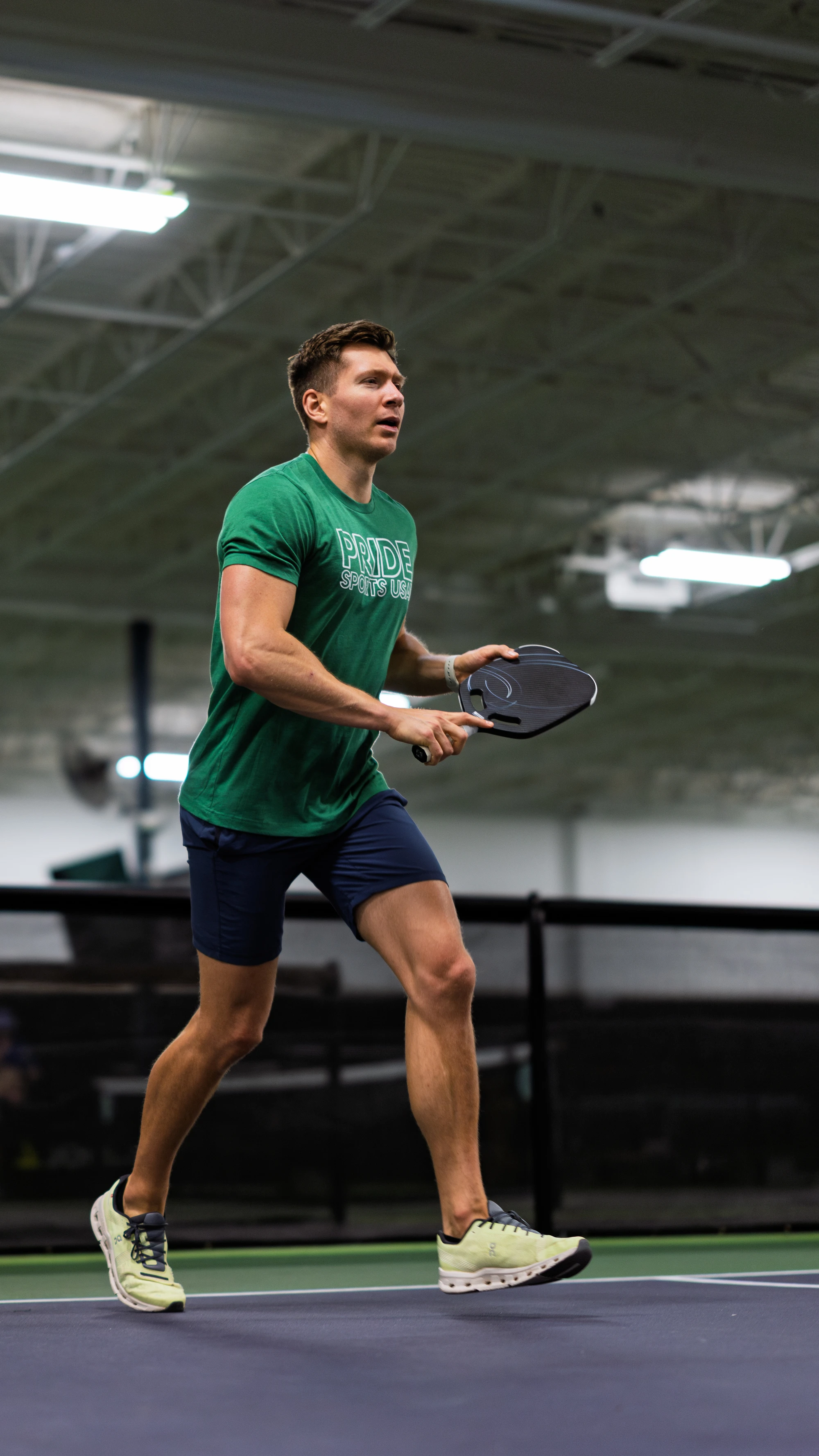 A pickleball player wearing a green "Pride Sports League" t-shirt and blue shorts prepares to hit a ball on an indoor court, holding a paddle while in motion.